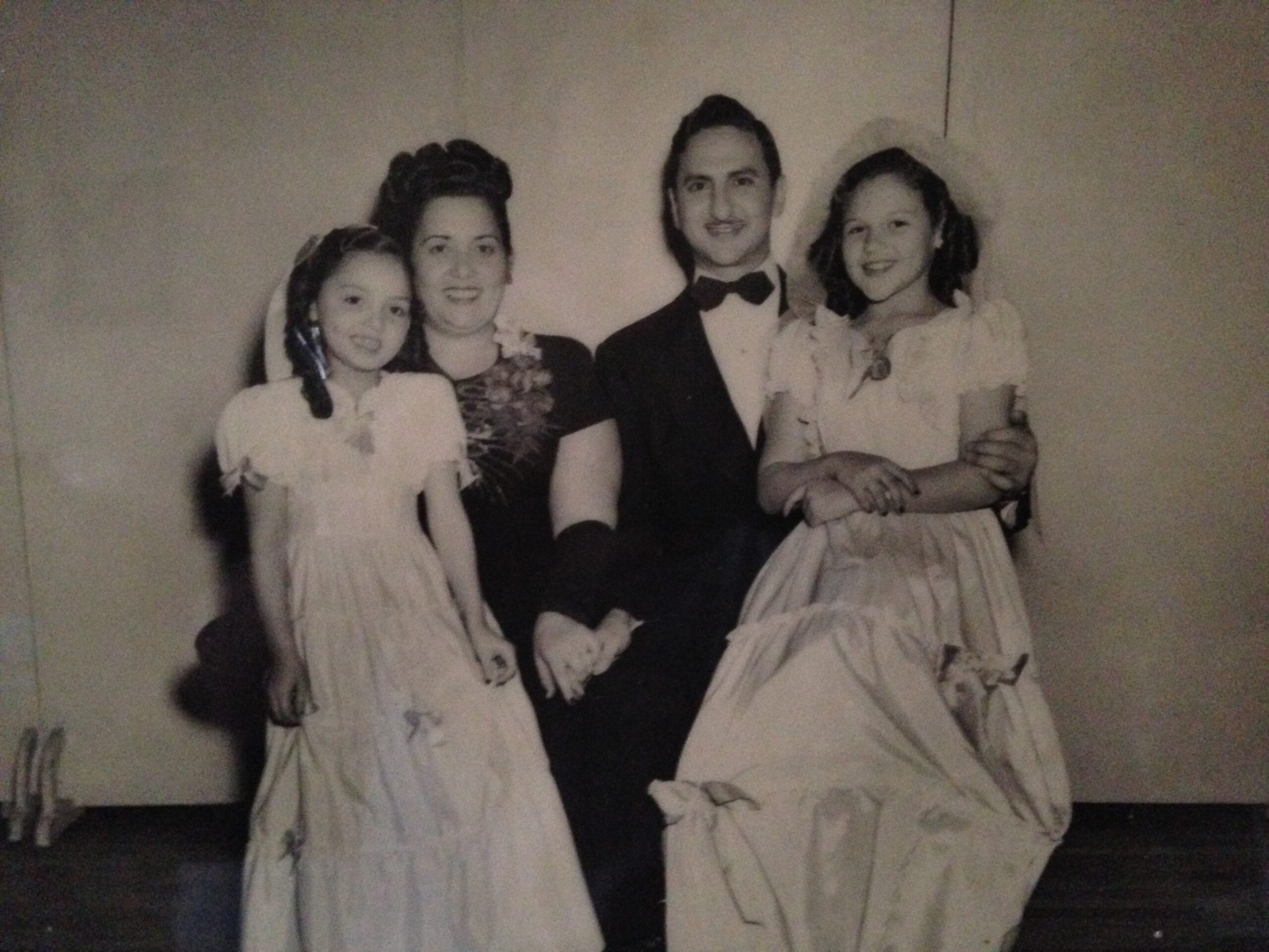 A black and white photo of a man and woman seated, in formal attire, with two young girls in long white dresses. Each girl is seated on the lap of either the man or the woman.