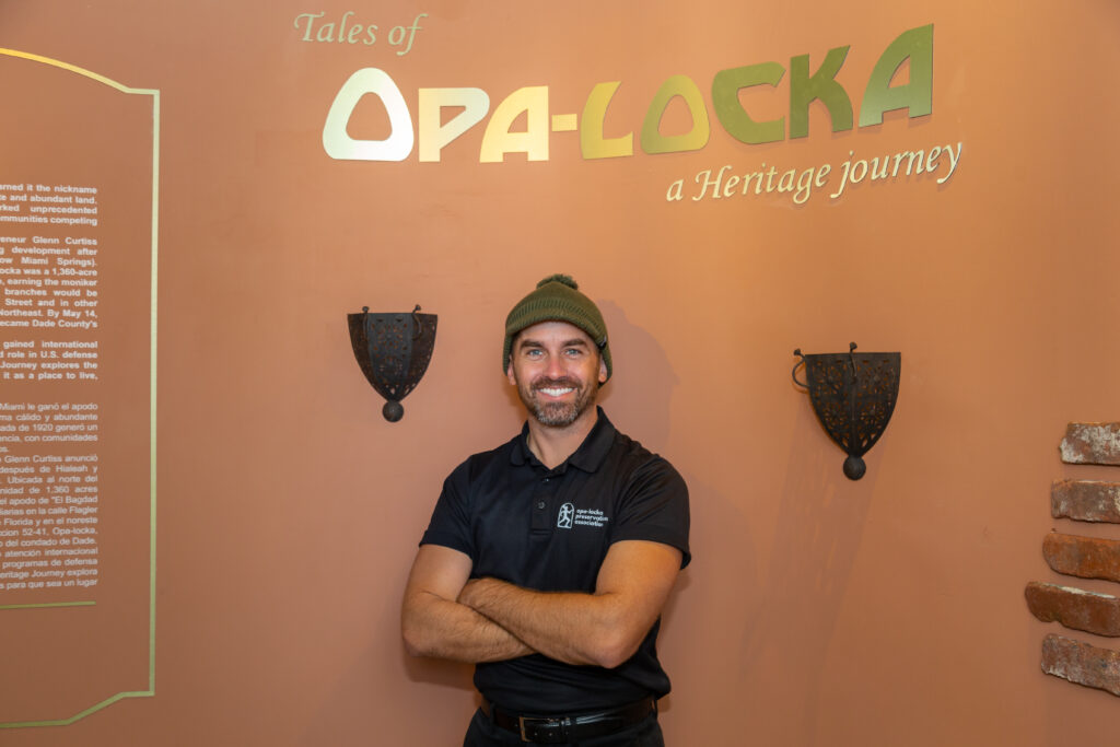 A person stands against a pink background under a sign that reads "Opa-locka." They wear a dark colored beanie, and navy polo shirt with their arms crossed as they smile towards the camera.