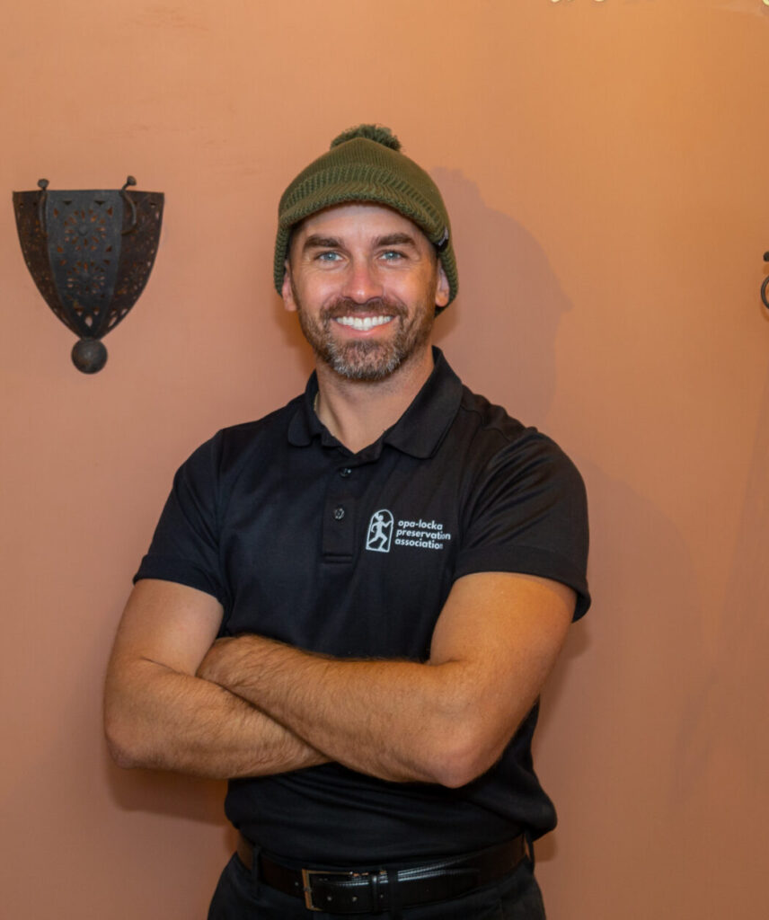 A person stands against a pink background under a sign that reads "Opa-locka." They wear a dark colored beanie, and navy polo shirt with their arms crossed as they smile towards the camera.