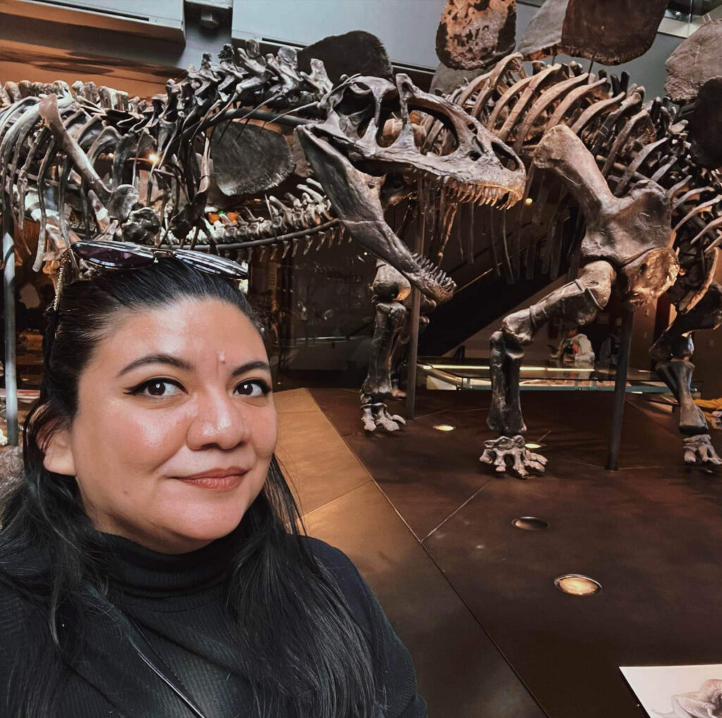 A woman stands in front of a museum exhibition with a fossilized dinosaur skeleton on view.