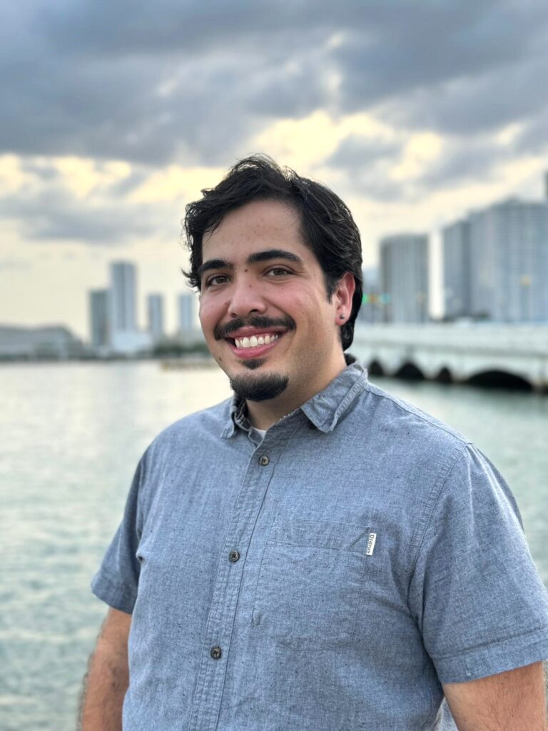 A person with short dark brown hair, a mustache, and short beard smiles towards the camera. They wear a short sleeve blue button down and stand in front of a body of water with a city skyline in the background.