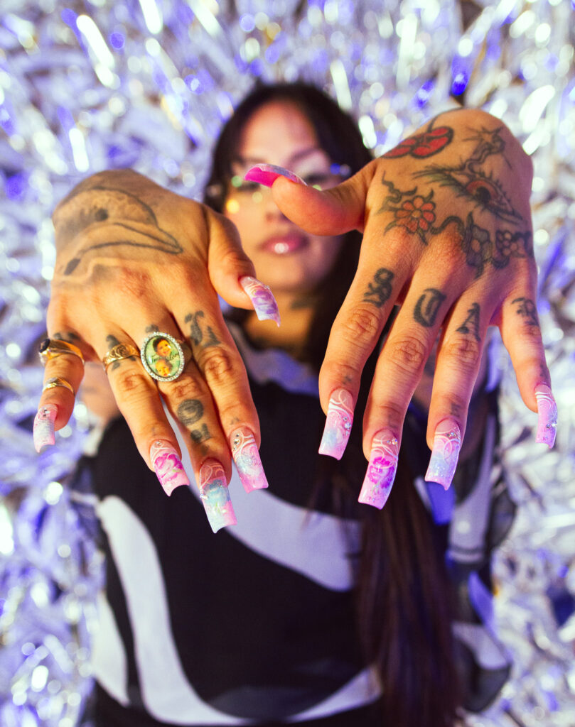 Kro holds out her hands in front of the camera with the focus on the purple. pink, and blue design of her nails, her jewelery, and the tattoos on her fingers and the back of her hands