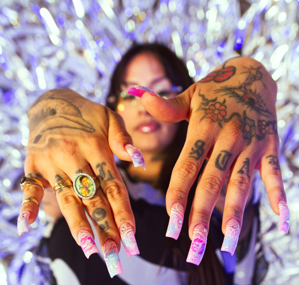 Kro holds out her hands in front of the camera with the focus on the purple. pink, and blue design of her nails, her jewelery, and the tattoos on her fingers and the back of her hands