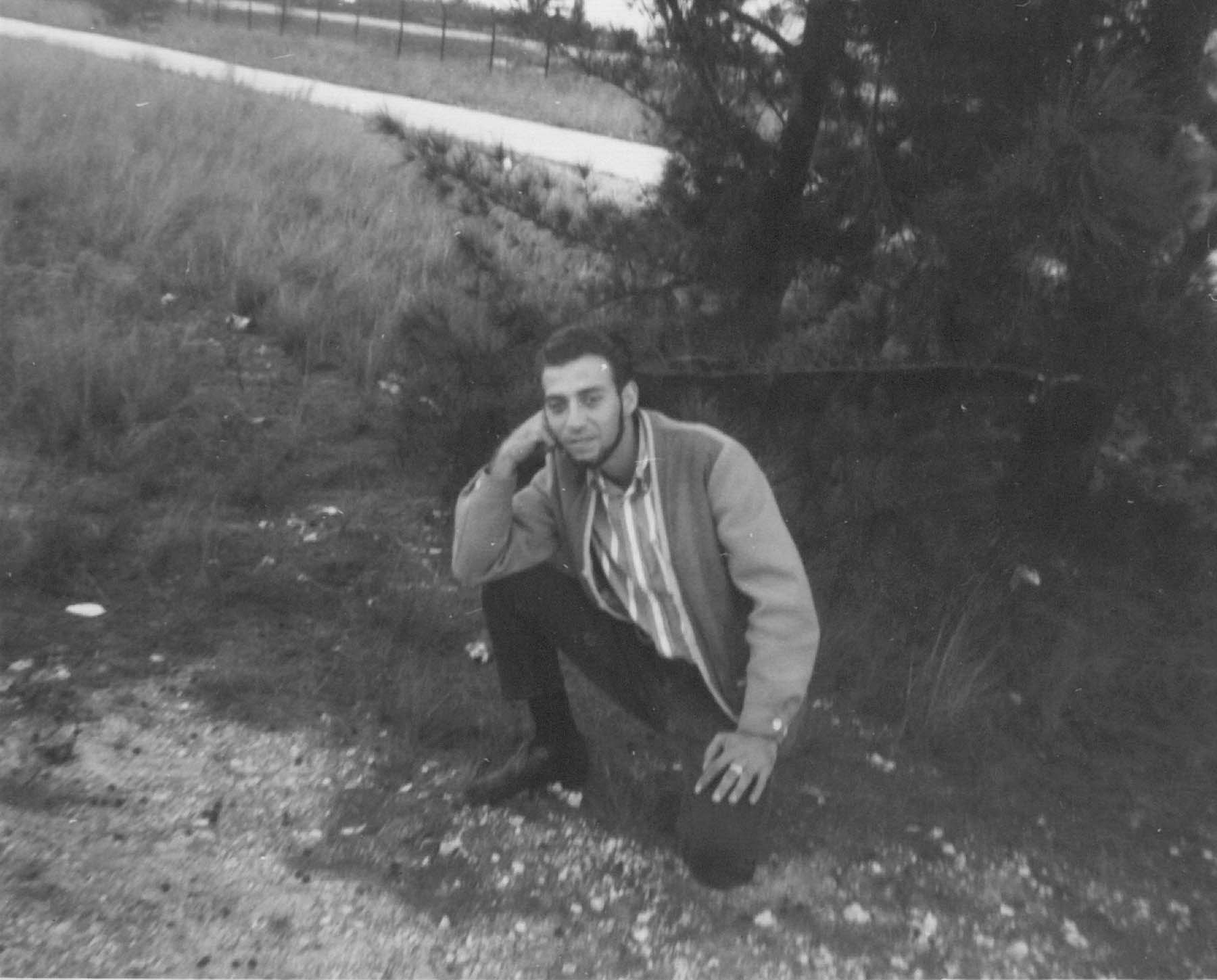 Black and white photo of a young man, Les Haber, kneeling in the grass by the road in 1964.