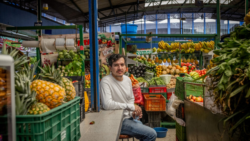 A person wearing a long sleeve white shirt and blue jeans sits amongst colorful crates of produce in an open-air market. They have short dark hair with a curl in the front and a short beard.