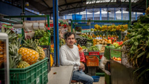 A person wearing a long sleeve white shirt and blue jeans sits amongst colorful crates of produce in an open-air market. They have short dark hair with a curl in the front and a short beard.