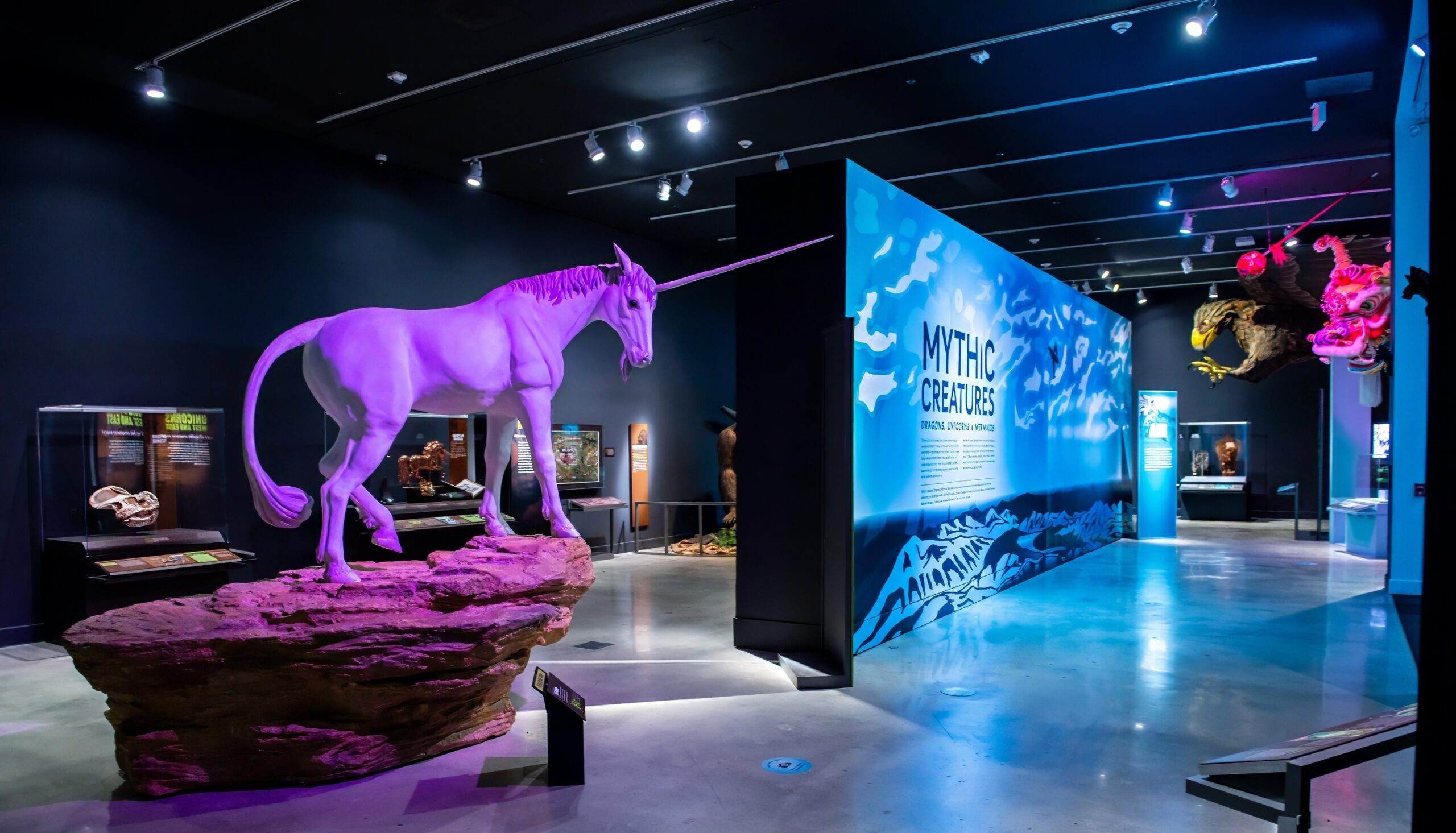 View of the Mythic Creatures exhibition; a unicorn sculpture stands atop a rock and is bathed in purple light. A blue and white entry panel with the text "Mythic Creatures" stands in the background.