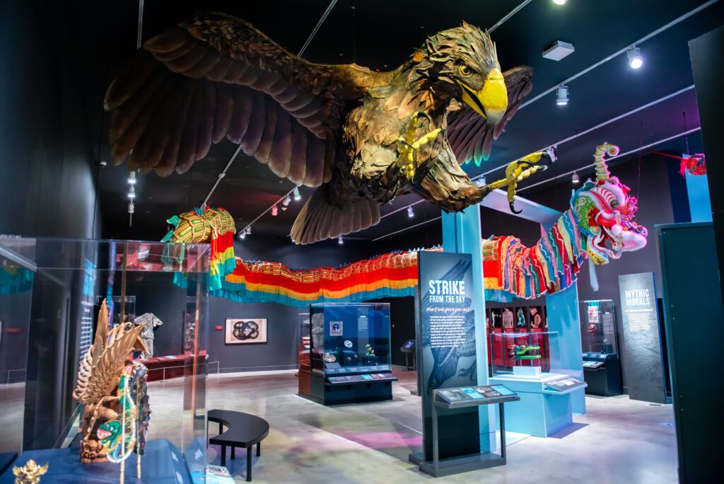 View of the Mythic Creatures museum exhibition. Exhibition infographic panels are standing throughout the exhibit and a large scale model of a brown bird with a yellow beak hangs from the ceiling. A colorful chinese parade dragon can be seen in the background hanging from the ceiling.