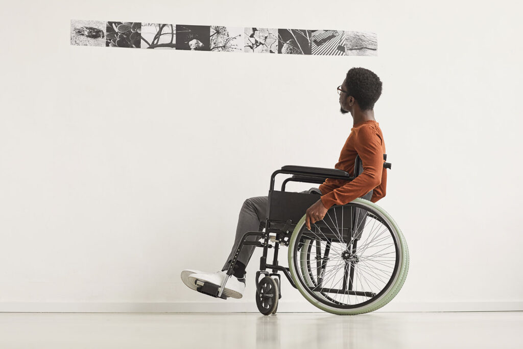 A person wearing an orange shirt, grey pants, and white sneakers who is seated in a wheelchair looking at black and white photographs displayed against a white wall.