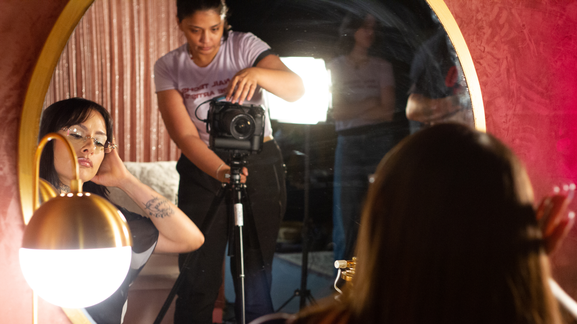 In the reflection of a large mirror, a person with long hair and glasses looks down while a photographer with a camera looks on.
