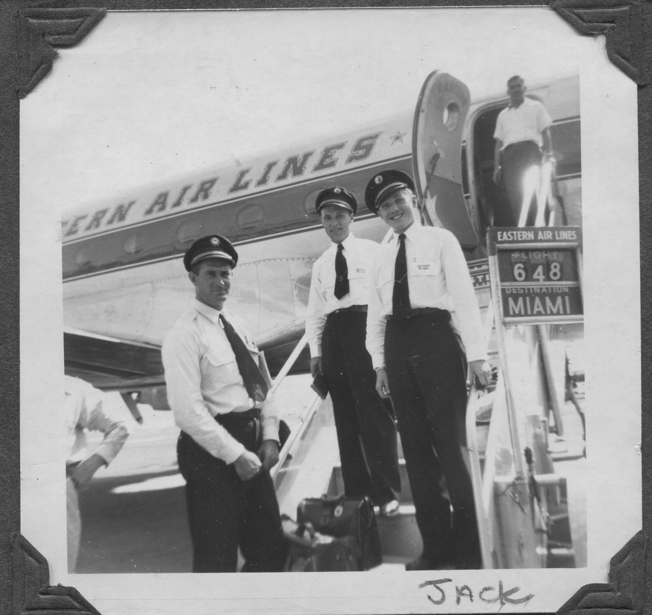 A black and white photo of three men in Eastern Airline Flight attendant uniforms standing in front of a plane at Miami International Airport, 1940s.