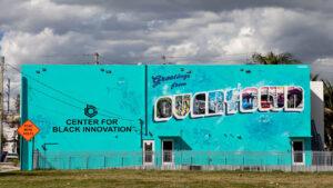 View of a mural in Overtown on a side of a building. The mural includes the words, "Center for Black Innovation" in black lettering and "Greetings from Overtown." Depictions of local sites and people are included inside the lettering of the latter phrase.