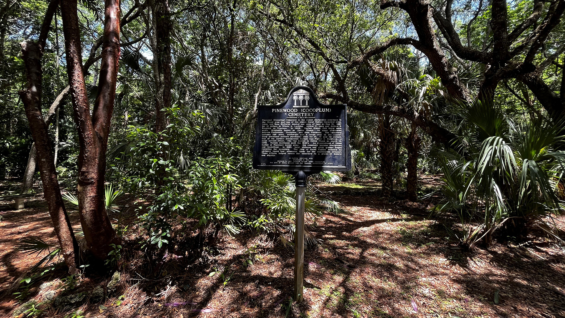 View of a tree covered cemetery with mulch covering the ground. A black historical marker with the words "Pinewood (Cocoplum) Cemetery)" stands in the center.
