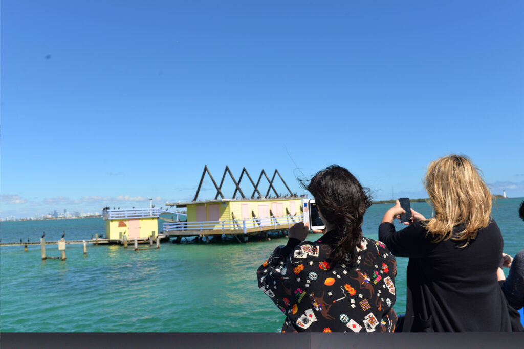 Two adults on a boat taking pictures of the yellow and pink Stiltsville building