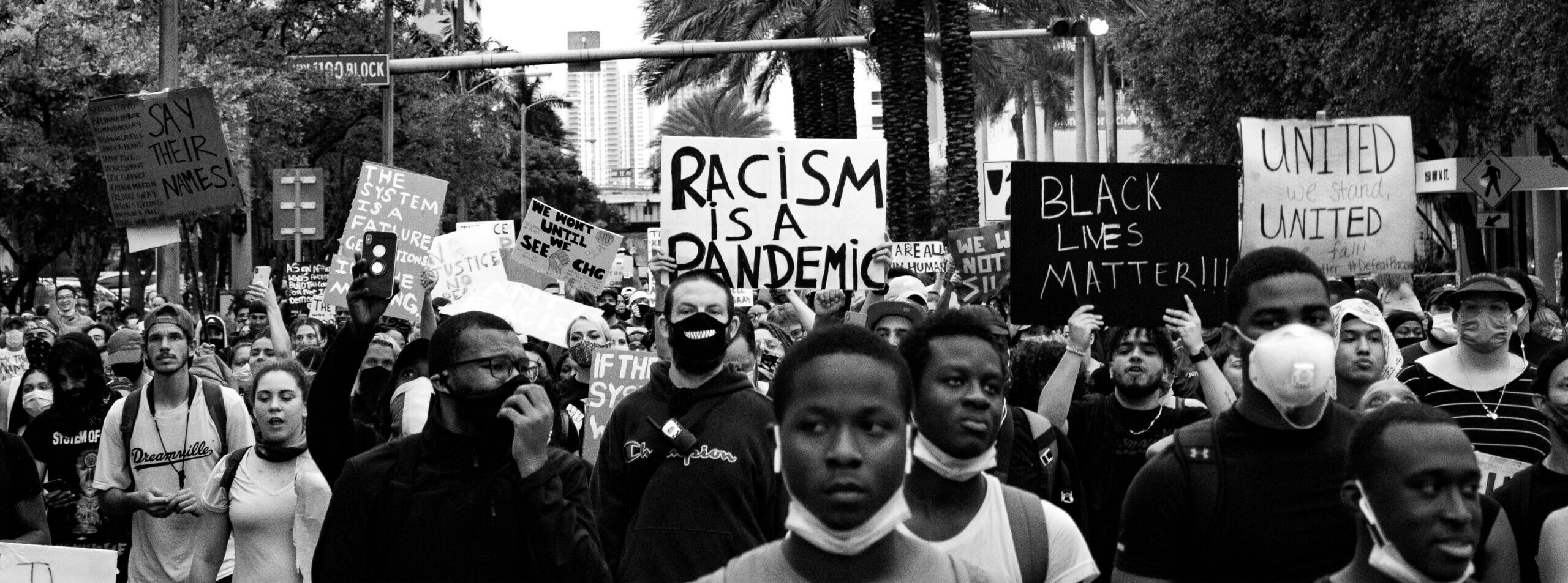 Black and white photograph of protestors in Downtown Miami. One holds a sign that reads, "racism is a pandemic."
