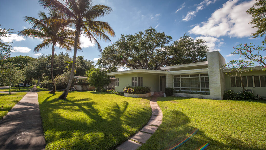 View of a one-level home in Miami Shores. The house sits on a green lawn with two palm trees against a blue sky. Courtesy of GMCVB/miamiandbeaches.com