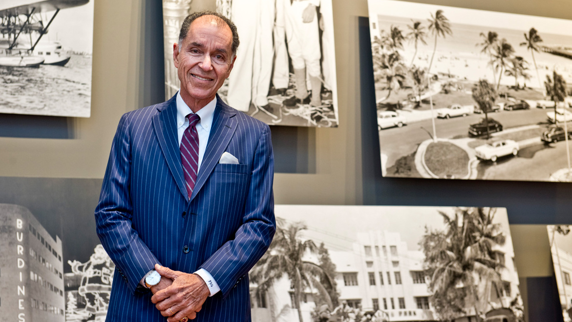 Dr. George, wearing a blue pinstripe suit, burgundy striped tie, and white shirt stands in front of a gallery of black and white archival images against a gray wall.