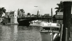 Black and white historical image of the Miami River. A boat is docked alongside the river with a bridge just behind it. In the background, a city skyline can be seen.