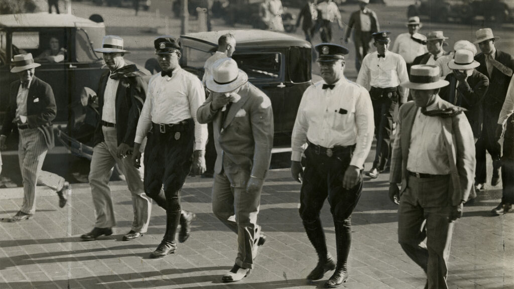 Six people walk in a row. The person in the center wears a hat and looks down while two police officers walk on either side.