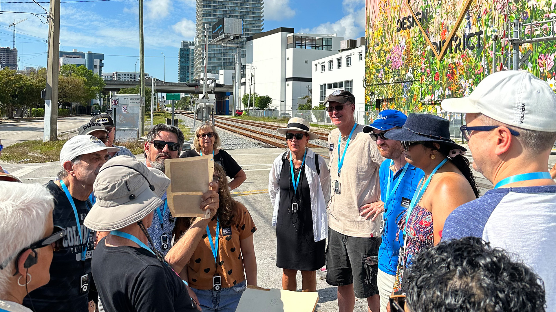A group stands on the side of a paved road in the Design District. They look towards a tour guide who stands wearing a hat, black shirt, and shorts and is holding a piece of paper up to the group.