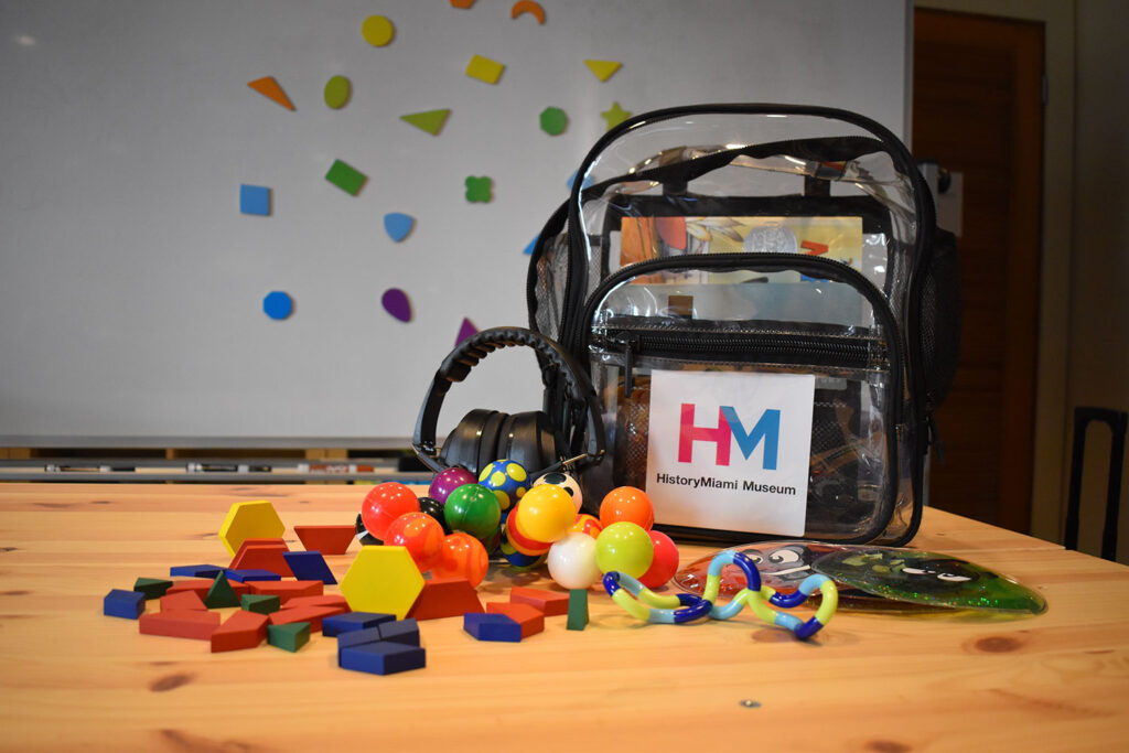 A clear and black outlined backpack sits on a wooden table in front of a white dry erase board with colorful magnets. On the table, colorful wooden geometric shapes and fidget toys sit in front of the backpack next to noise reducing headphones.
