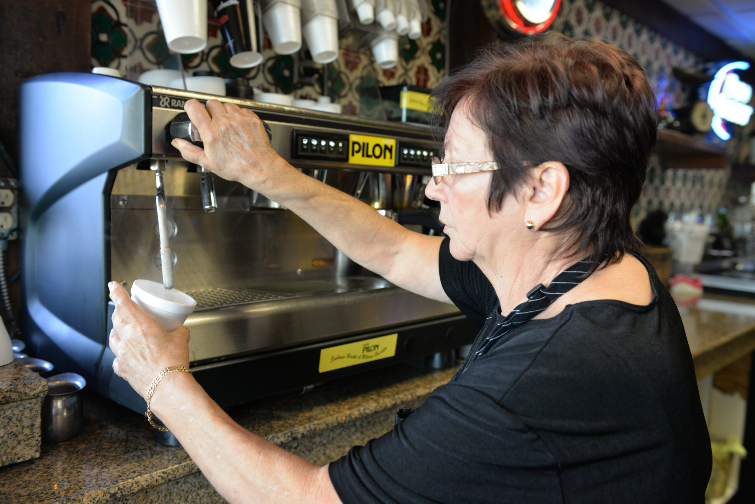 Iris Diaz turns a knob on an espresso machine to froth the milk for a "cafe con leche" she holds in her other hand