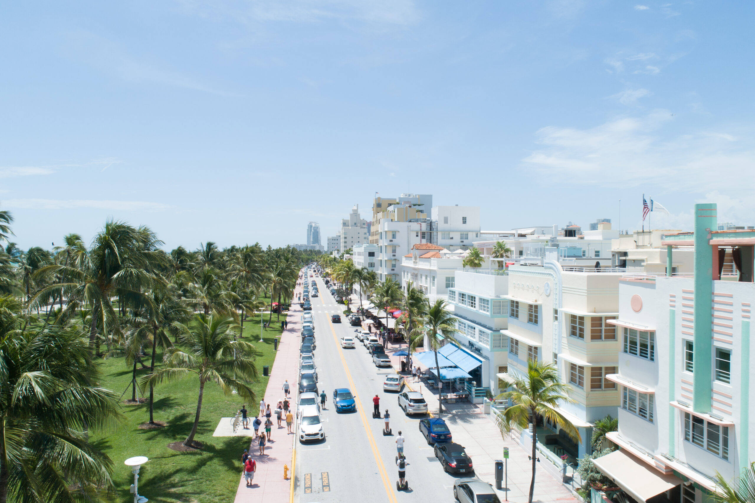 Aerial view of South Beach off of Ocean Drive. To the left, a linear grassy park is seen dotted with palm trees next to a paved street. Along the other side of the street, colorful art deco buildings are seen.