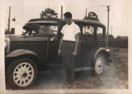 A black and white photo of a man, Tom Harrington, with his first car, a 1931 Buick. A small black dog is on the hood of the car.
