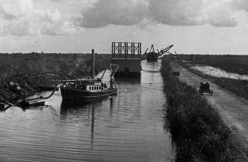 A dredge dug fill for the Tamiami Trail roadbed, and dug Tamiami Canal. This photo shows, from front to back, a service boat, drill barge, and dredge in Tamiami Canal, 1926 or 1927. The dredge and roadbed became an unintentional dam, impeding the flow of water from the north to the south. X-1311-1