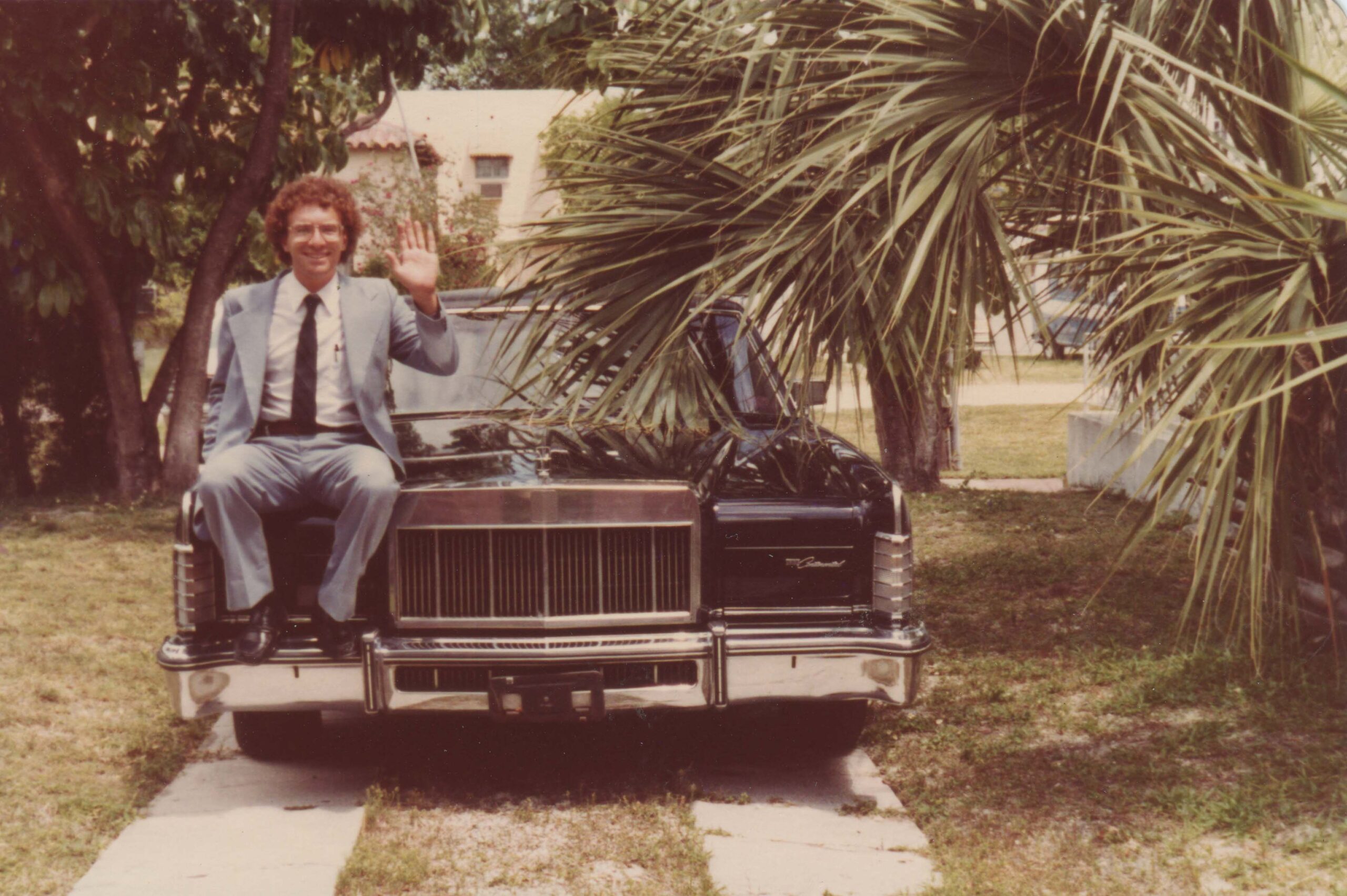 A man in a suit, Zeb Beitchman, suits on the hood of a car and waves. Photo taken in 1983.