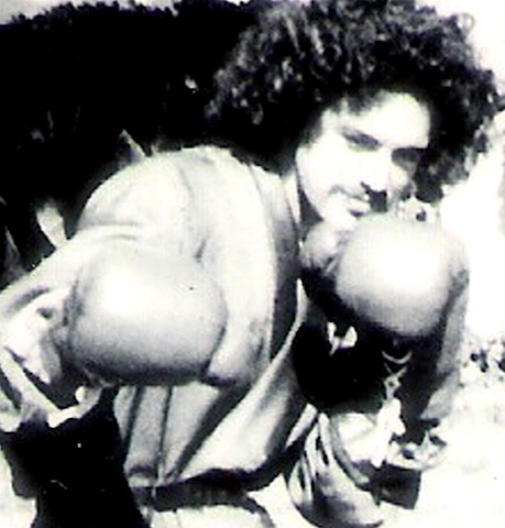 A black and white photo of a young man wearing boxing gloves with his hands by his face.