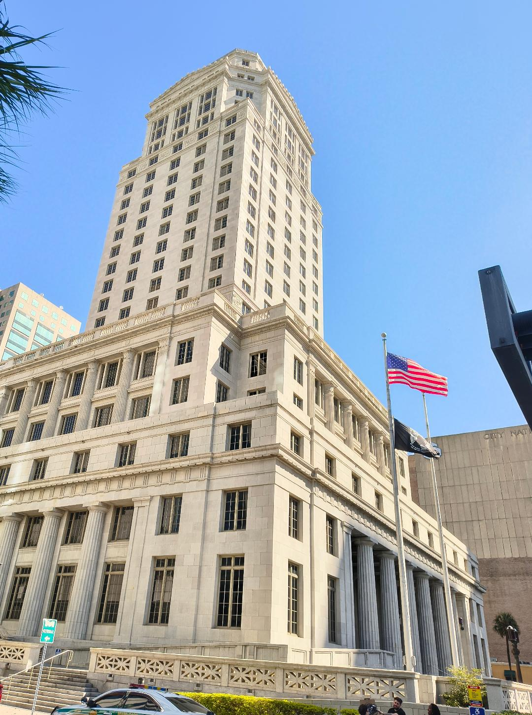 Street view of the Miami Dade County Courthouse, a tall beige building with a tall central tower. There are steps and a long ramp in front of the building and an American flag and POW flag fly on a flagpole on the corner.