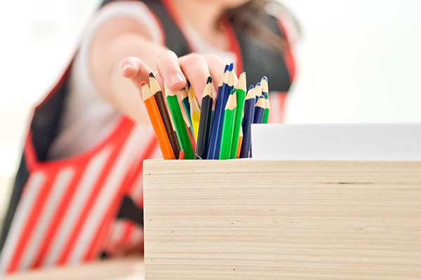 A child's hand reaching for standing colored pencils in a wooden container