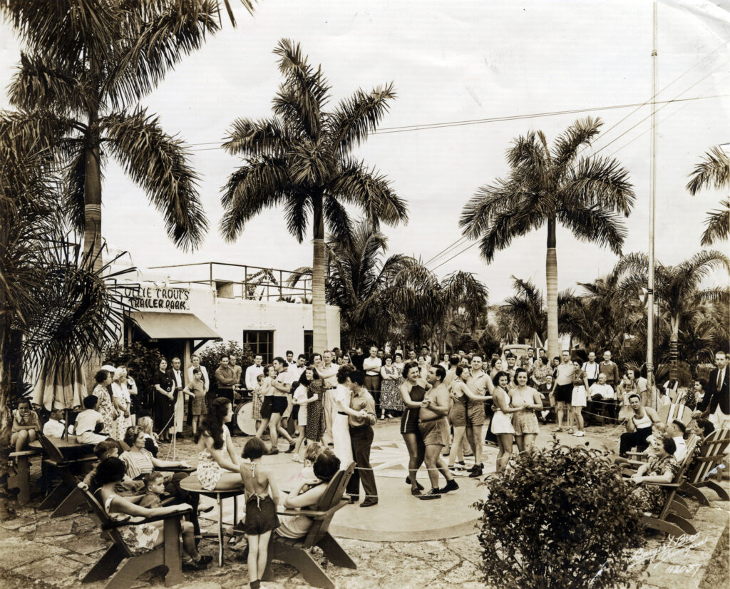 A sepia image of people dancing on a dance floor as some look on from patio chairs outdoors in front of a building with the sign "Ollie Trout's Trailer Park" above an awning. 
