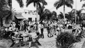 A sepia image of people dancing on a dance floor as some look on from patio chairs outdoors in front of a building with the sign "Ollie Trout's Trailer Park" above an awning.