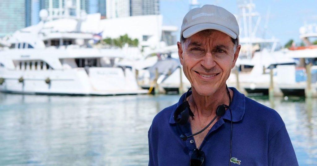 Dr. George wears a blue polo shirt and beige hat standing in front of a marina with boats in the distance.