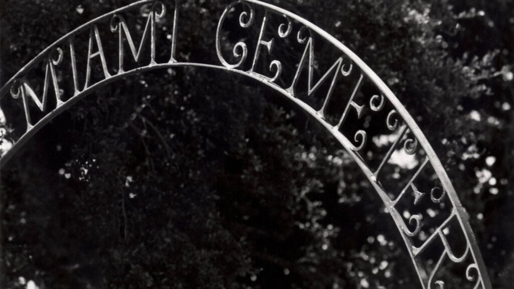 Black and white image of Miami City Cemetery entrance sign; metalwork script with "Miami Cemetery" can be seen in the center of the image with trees in the background.