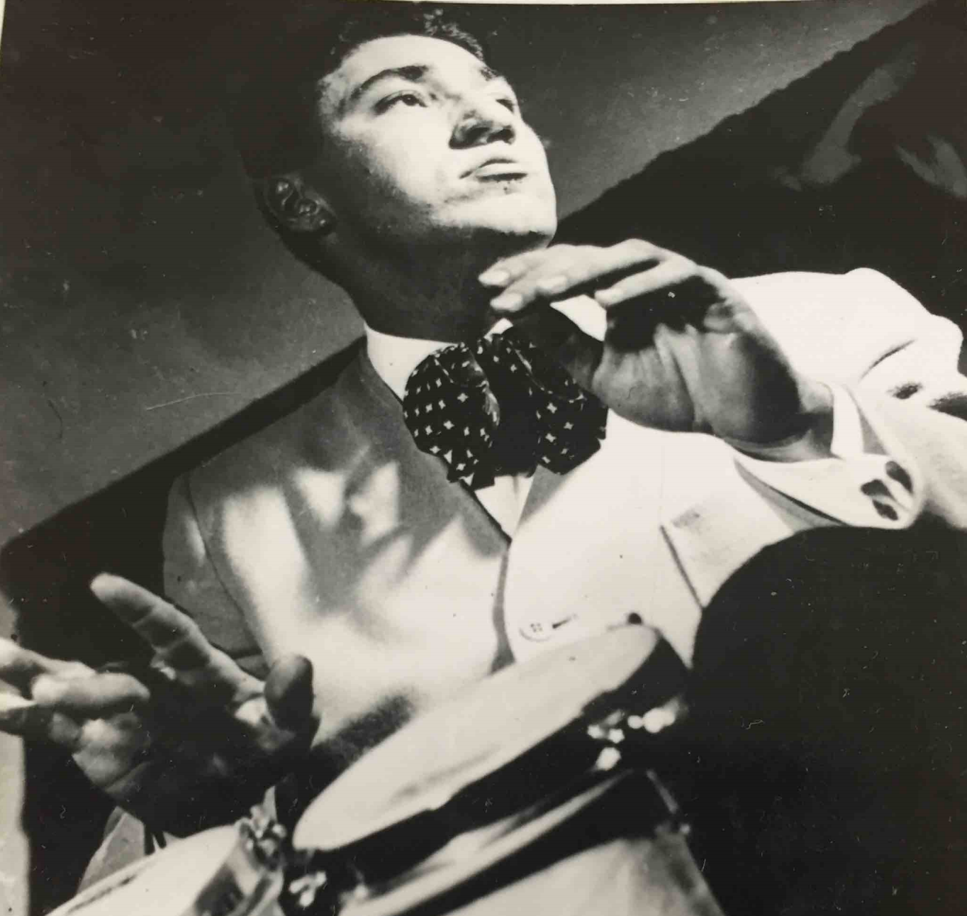 A black and white 1950s photo of a man playing bongos. He's wearing a suit and looking off camera.