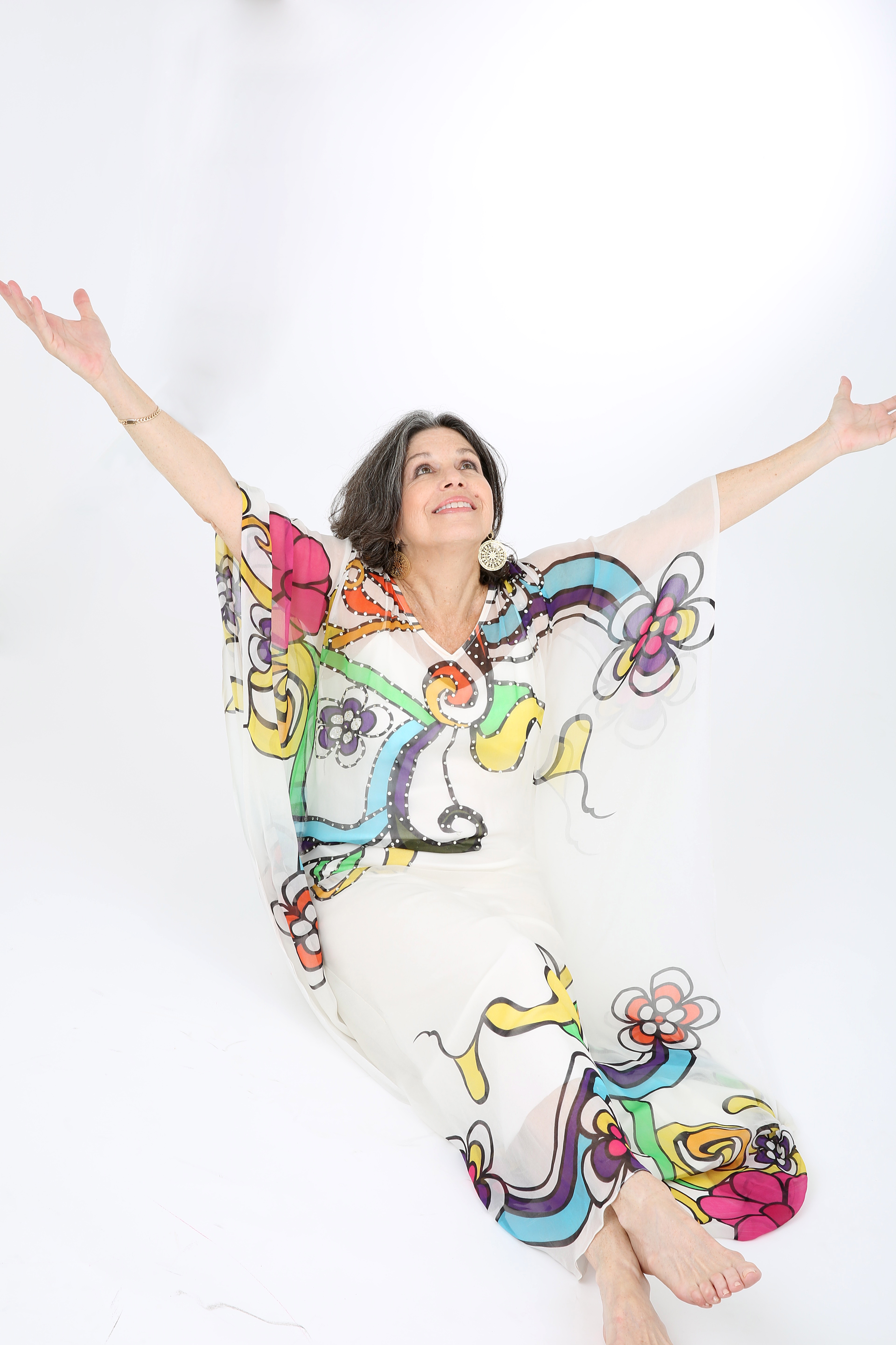 Susana Behar Levy is wearing a colorful dress against a white background and she has her arms thrown up in the air.