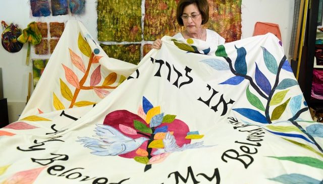 Nancy holds the edge of the chuppah in her arms as it is laid out over a table. The center has two birds holding a branch with colored leaves in their beaks with a red heart behind them. Under the heart it reads, 