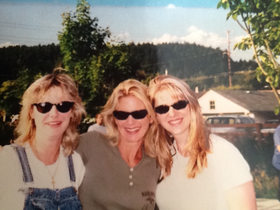 A close-up photo of three blonde women in sunglasses. The woman in the middle has her arms over the shoulders of the other two.
