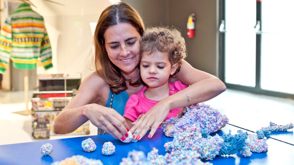 A person with a young child in their lap sit at a blue table in a museum exhibition. They interact with a foam material made of smaller colorful foam beads.