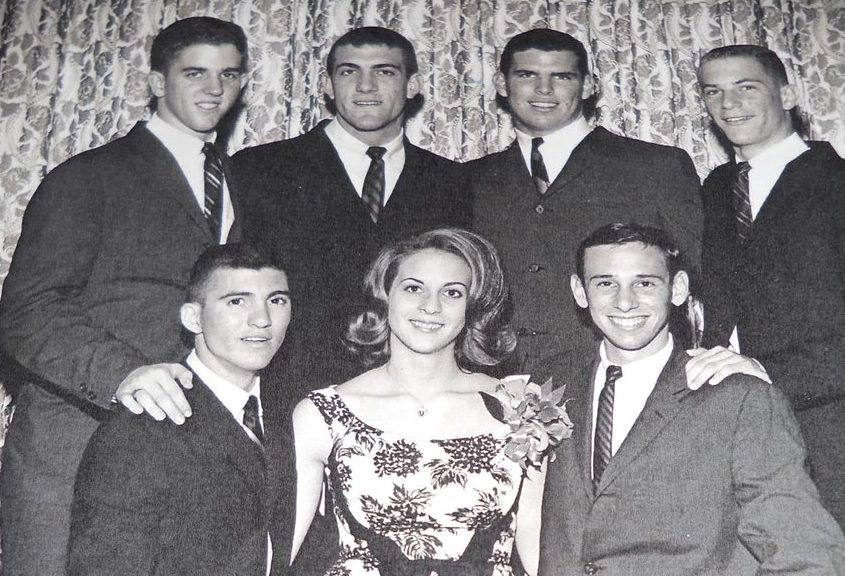 A black and white photo of seven young people in formal attire. In the back row are four young men standing and in the front is a woman seated with two men kneeling at either side. The storyteller, Orin Hollander is the young man in the front row on the left. This was taken at the Sigma Rho Formal in 1963.